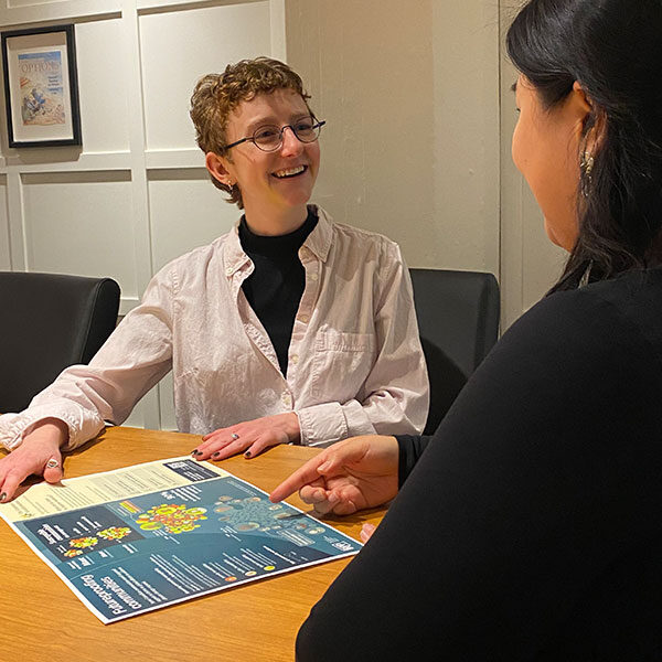 EN: Two people in conversation; one wears a white coat and smiles while pointing to a colourful diagram. FR: Deux personnes en conversation; l’une porte un sarrau blanc et sourit en montrant un diagramme coloré.
