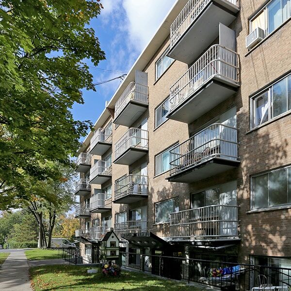 EN: A modern multi-storey apartment building with balconies, surrounded by trees and green grass. FR: Un immeuble résidentiel moderne à plusieurs étages avec balcons, entouré d'arbres et de pelouse.