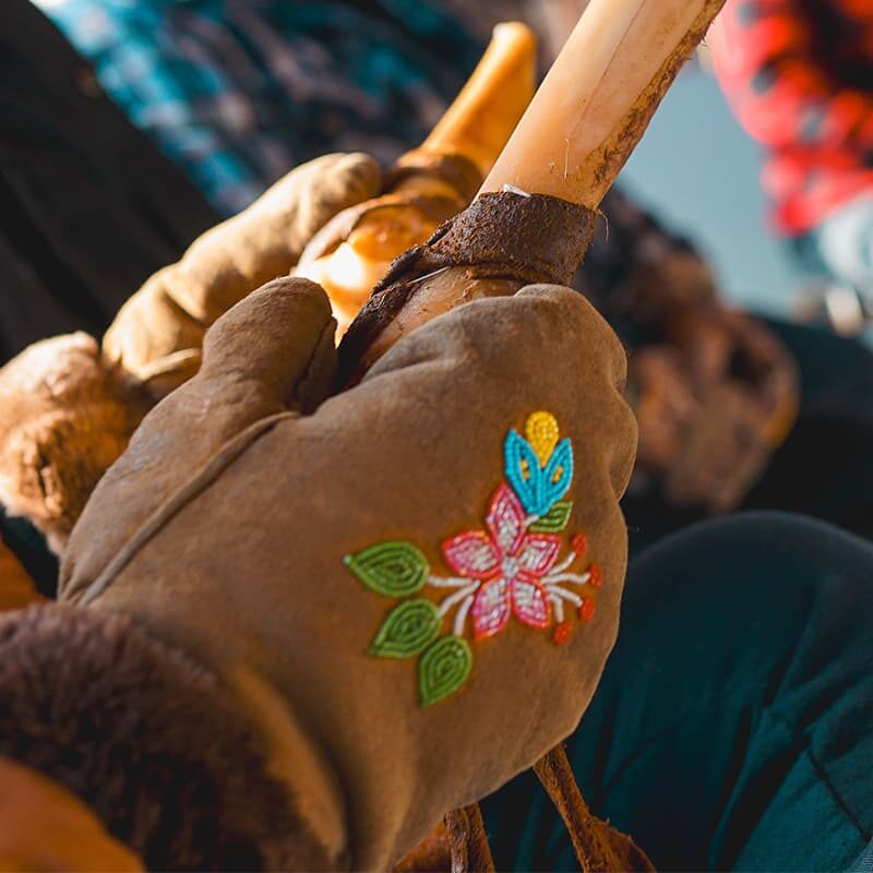 EN: A close-up of a hand wearing a glove decorated with a beaded floral design, holding a carved piece of bone. Several people sit in a semi-circle in the background, slightly out of focus, suggesting a group gathering or cultural activity. FR: Gros plan d'une main portant un gant orné d'une fleur perlée, tenant un morceau d'os sculpté. Plusieurs personnes sont assises en demi-cercle à l’arrière-plan, légèrement floues, suggérant un rassemblement ou une activité culturelle.